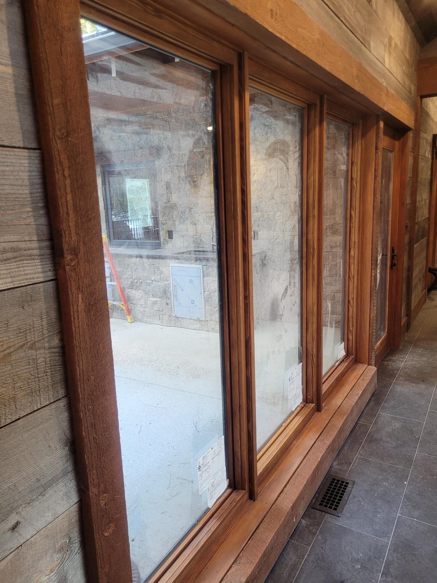 Interior view through wood-framed windows to a stone outdoor kitchen and patio